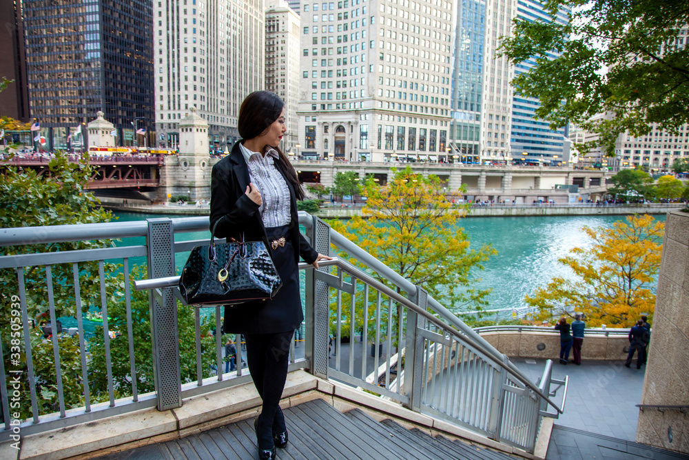 Naklejka premium Beautiful Chinese Asian woman in fashionable business attire is enjoying a nice day walking around downtown Chicago in the afternoon. She carries a trendy handbag and black blazer with red lipstick
