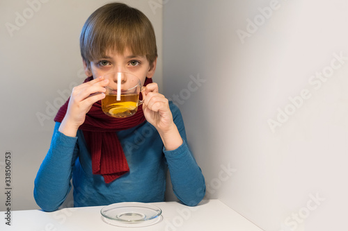 Caucasian boy in a warm sweater and scarf drinks hot tea with lemon. The baby looks into the camera. Concept of catarrhal diseases. Copyspace
