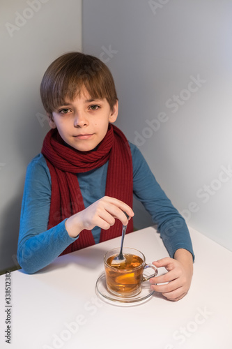 Caucasian boy in a warm sweater and scarf drinks hot tea with lemon. The baby looks into the camera. Concept of catarrhal diseases. Copyspace