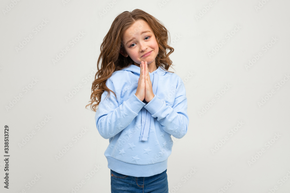 cute teenage girl in a blue hoodie joyfully smiles asks holding her hands folded against a light gray background cute teenage girl in a blue hoodie joyfully smiles asks holding her hands folded against a light gray background