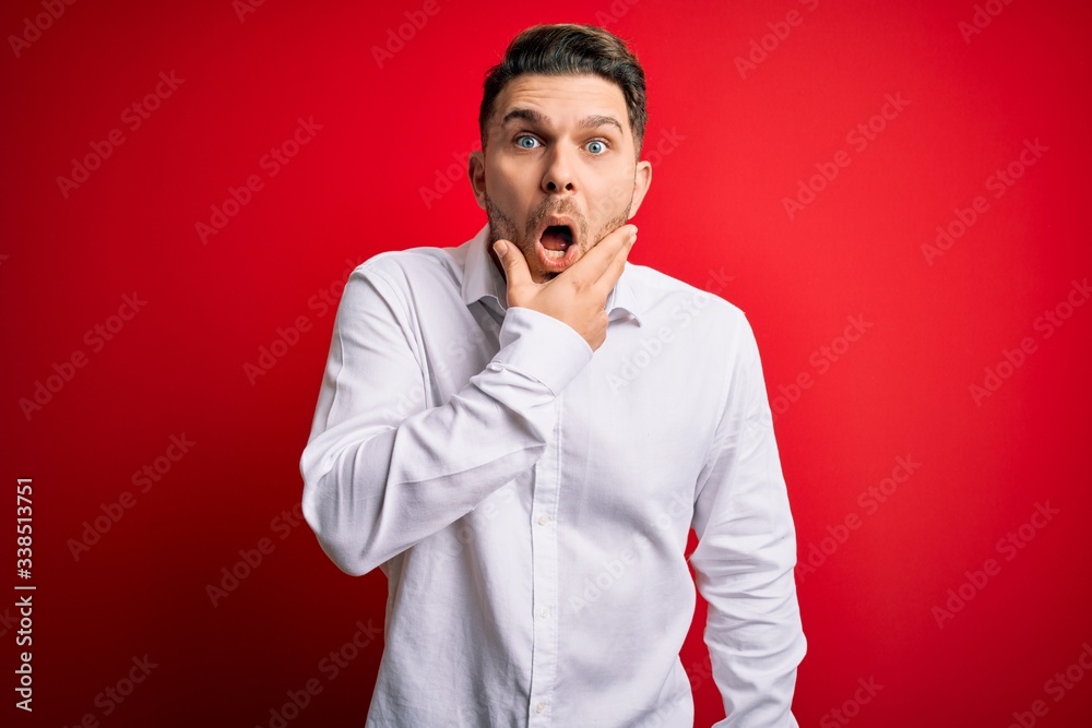 Young business man with blue eyes wearing elegant shirt standing over red isolated background Looking fascinated with disbelief, surprise and amazed expression with hands on chin