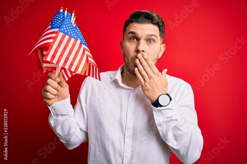 Young man with blue eyes holding flag of united states of america over red isolated background cover mouth with hand shocked with shame for mistake, expression of fear, scared in silence, secret