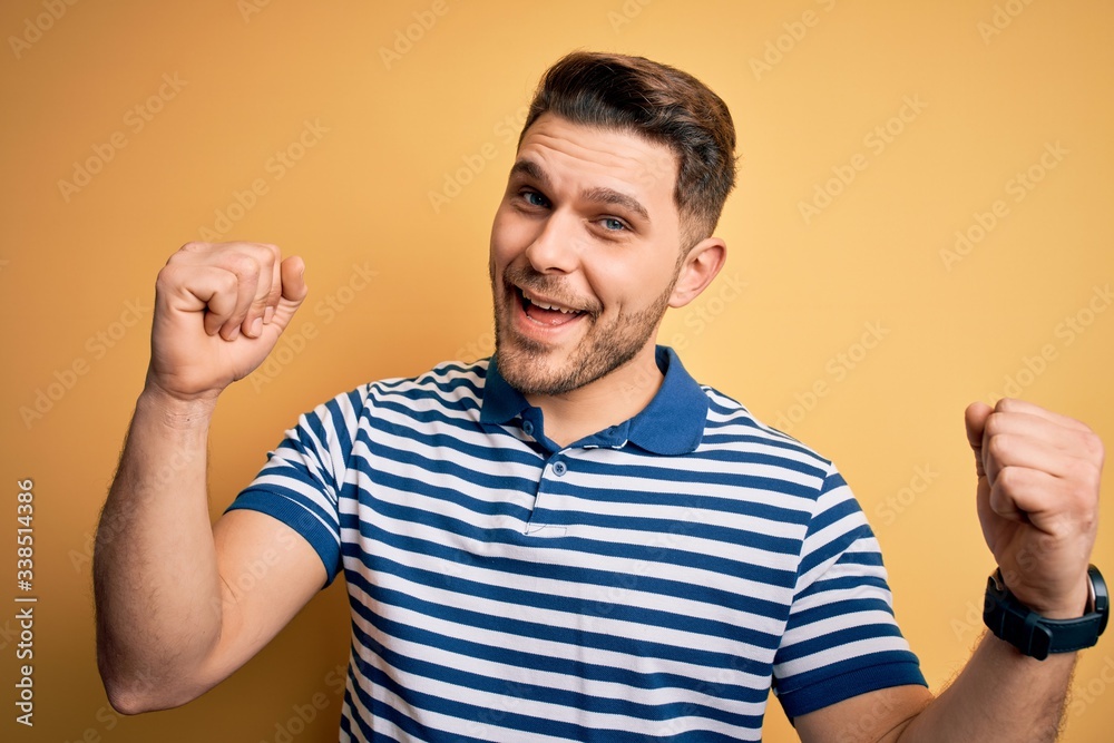 Young man with blue eyes wearing casual striped t-shirt over yellow background celebrating surprised and amazed for success with arms raised and open eyes. Winner concept. Young man with blue eyes wearing casual striped t-shirt over yellow background celebrating surprised and amazed for success with arms raised and open eyes. Winner concept.