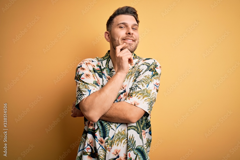 Young man with blue eyes on vacation wearing floral summer shirt over yellow background looking confident at the camera with smile with crossed arms and hand raised on chin. Thinking positive. Young man with blue eyes on vacation wearing floral summer shirt over yellow background looking confident at the camera with smile with crossed arms and hand raised on chin. Thinking positive.