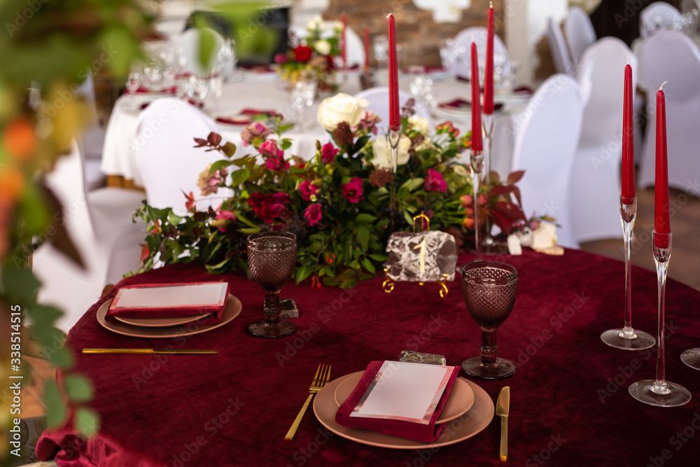 Place at the banquet table: the menu card lies on a red napkin on plates, a gilt fork and knife on the sides of the plate, a glass of brown glass, flower arrangement, red candles