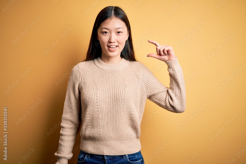 Young beautiful asian woman wearing casual sweater over yellow isolated background smiling and confident gesturing with hand doing small size sign with fingers looking and the camera. Measure concept. Young beautiful asian woman wearing casual sweater over yellow isolated background smiling and confident gesturing with hand doing small size sign with fingers looking and the camera. Measure concept.
