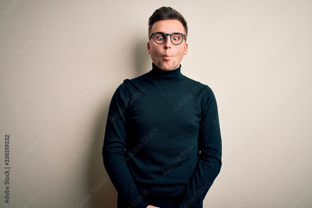 Young handsome caucasian man wearing glasses and casual sweater over isolated background making fish face with lips, crazy and comical gesture. Funny expression.
