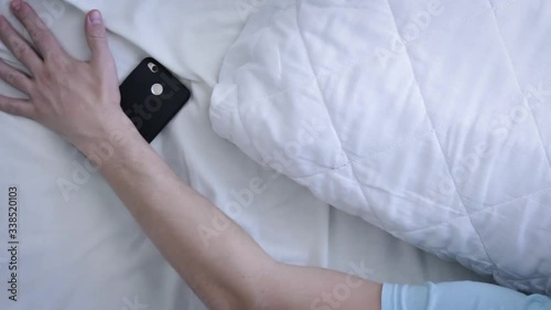 Young  man waked up, looks on phone and return to sleep again. Alarm Clock On Phone. Tired man Waking Up In Bed At Bedroom. Modern technologies concept