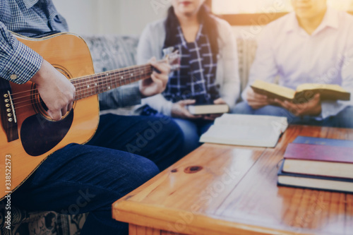 A young man is playing guitar and sings a song from Christian hymn book with his friends at home, Christian family worship concept