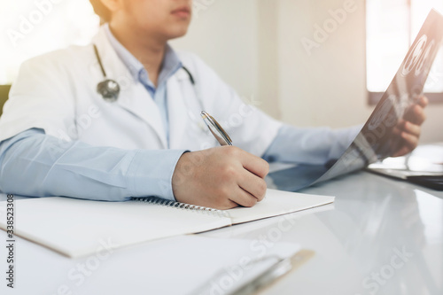 Young Asian doctor with Medical stethoscope holding X-ray film while writing on notebook in an office at the hospital
