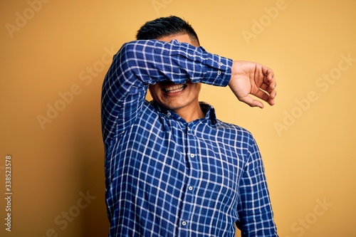 Young handsome latin man wearing casual shirt and glasses over yellow background covering eyes with arm smiling cheerful and funny. Blind concept.