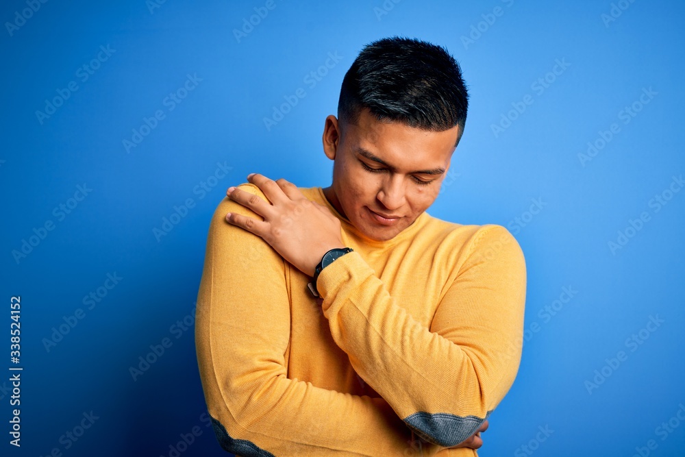 Young handsome latin man wearing yellow casual sweater over isolated blue background Hugging oneself happy and positive, smiling confident. Self love and self care Young handsome latin man wearing yellow casual sweater over isolated blue background Hugging oneself happy and positive, smiling confident. Self love and self care