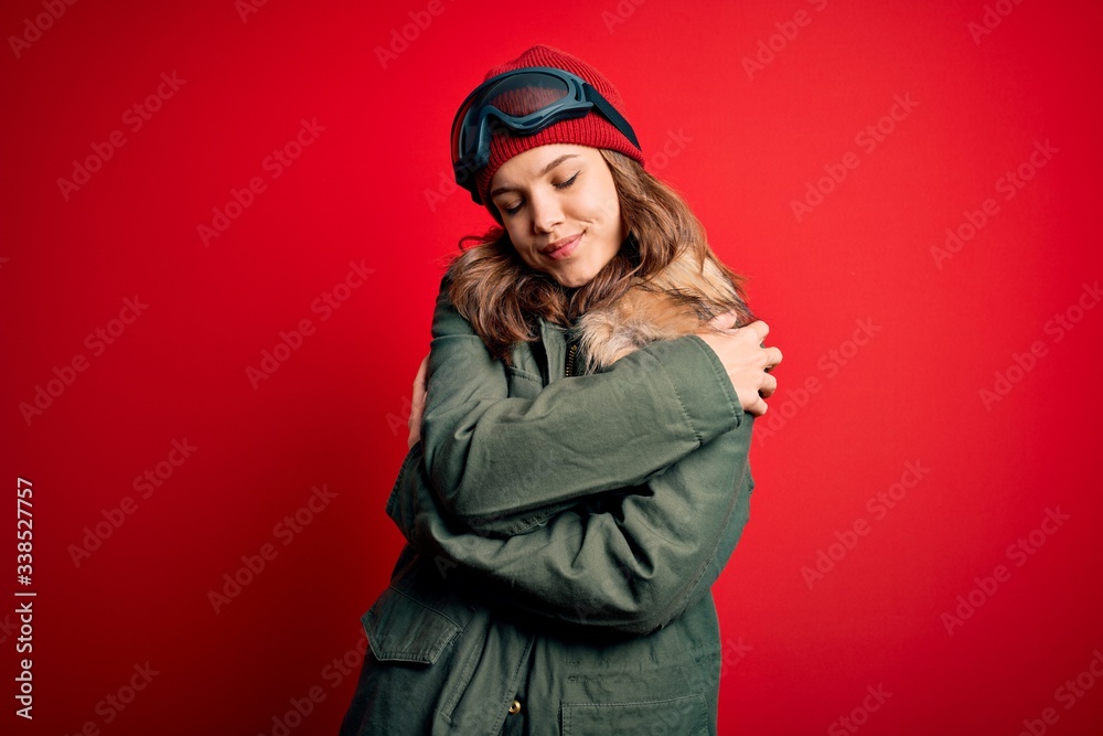 Young blonde girl wearing ski glasses and winter coat for ski weather over red background Hugging oneself happy and positive, smiling confident. Self love and self care Young blonde girl wearing ski glasses and winter coat for ski weather over red background Hugging oneself happy and positive, smiling confident. Self love and self care