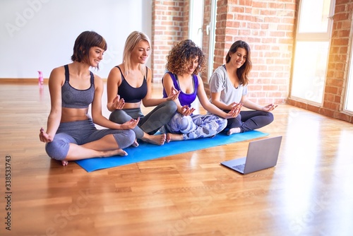 Young beautiful group of sportswomen smiling happy practicing yoga. Sitting doing lotus pose using laptop at gym