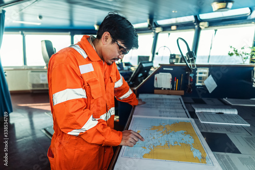 Filipino deck Officer on bridge of vessel or ship wearing coverall during navigaton watch at sea . He is plotting position on chart