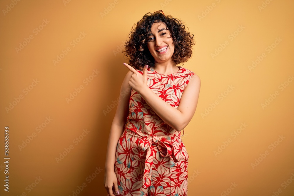 Young beautiful curly arab woman on vacation wearing summer floral dress and sunglasses cheerful with a smile of face pointing with hand and finger up to the side with happy and natural expression Young beautiful curly arab woman on vacation wearing summer floral dress and sunglasses cheerful with a smile of face pointing with hand and finger up to the side with happy and natural expression