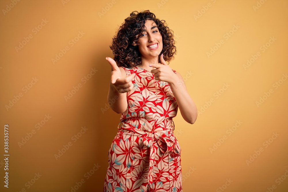 Young beautiful curly arab woman on vacation wearing summer floral dress and sunglasses pointing fingers to camera with happy and funny face. Good energy and vibes. Young beautiful curly arab woman on vacation wearing summer floral dress and sunglasses pointing fingers to camera with happy and funny face. Good energy and vibes.