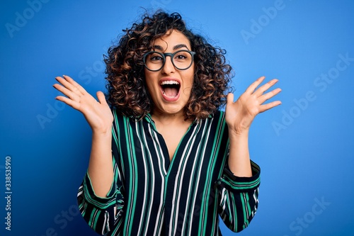 Young beautiful curly arab woman wearing striped shirt and glasses over blue background celebrating crazy and amazed for success with arms raised and open eyes screaming excited. Winner concept