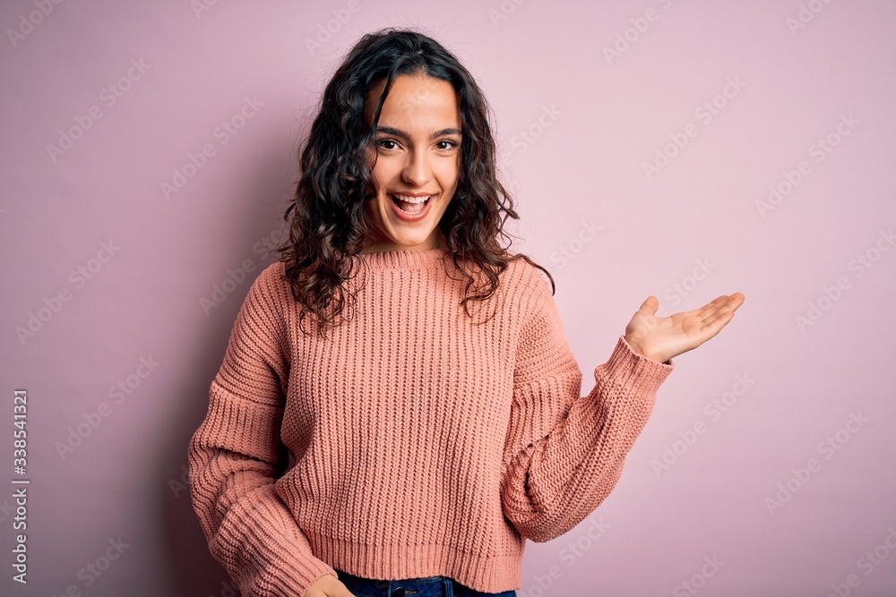 Young beautiful woman with curly hair wearing casual sweater over isolated pink background smiling cheerful presenting and pointing with palm of hand looking at the camera. Young beautiful woman with curly hair wearing casual sweater over isolated pink background smiling cheerful presenting and pointing with palm of hand looking at the camera.
