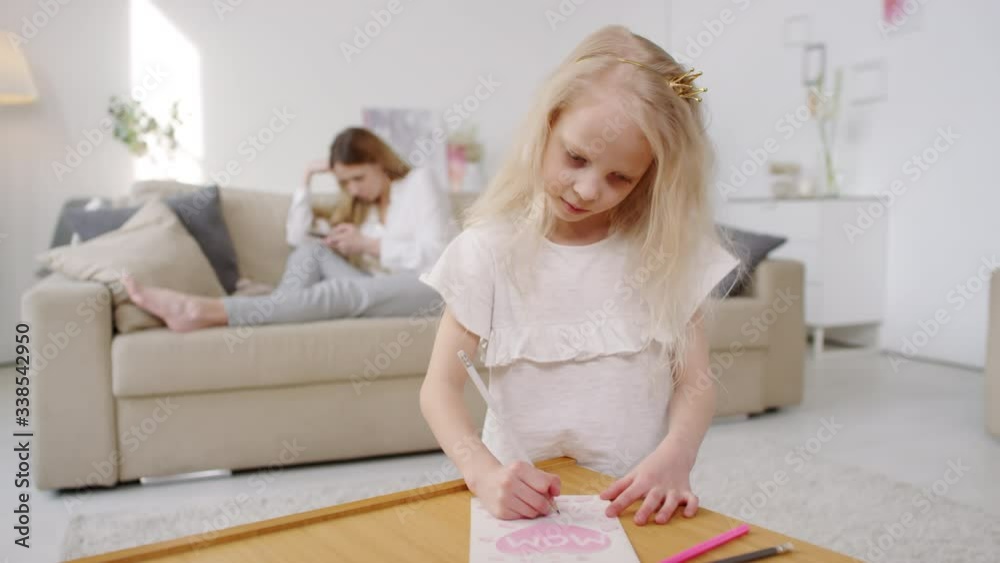 Medium shot of cute little girl wearing hairband with crown making mothers day postcard as her mother sitting on couch and looking at mobile phone Medium shot of cute little girl wearing hairband with crown making mothers day postcard as her mother sitting on couch and looking at mobile phone