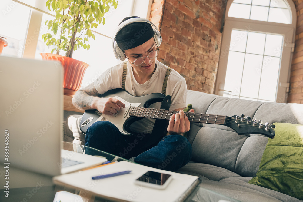 Man studying at home during online courses or free information by hisself. Becomes musician, guitarist while isolated, quarantine against coronavirus spreading. Using laptop, smartphone, headphones.