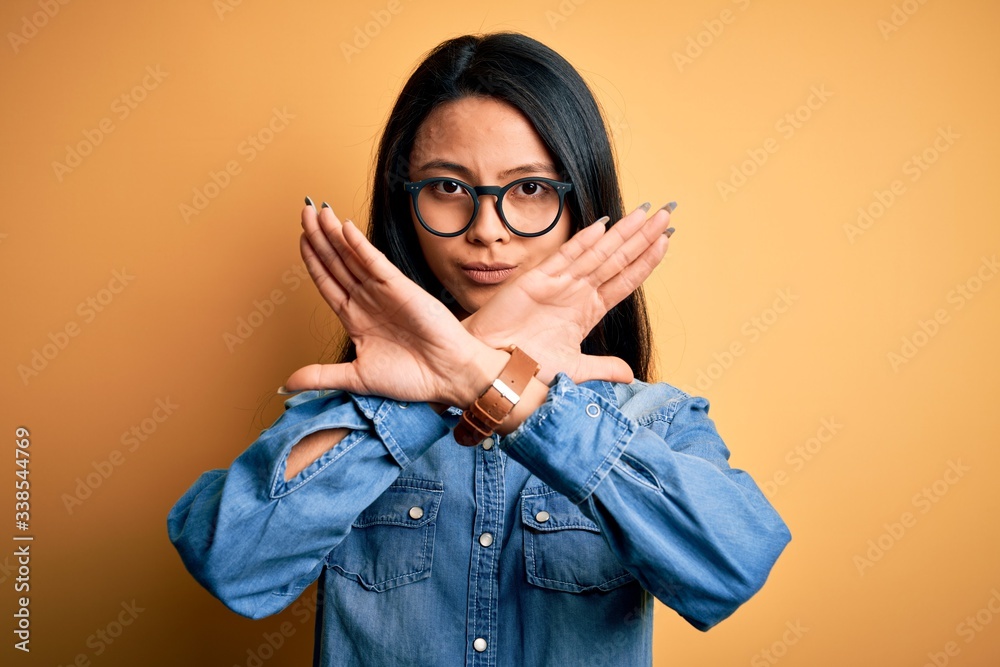 Young beautiful chinese woman wearing casual denim shirt over isolated yellow background Rejection expression crossing arms doing negative sign, angry face
