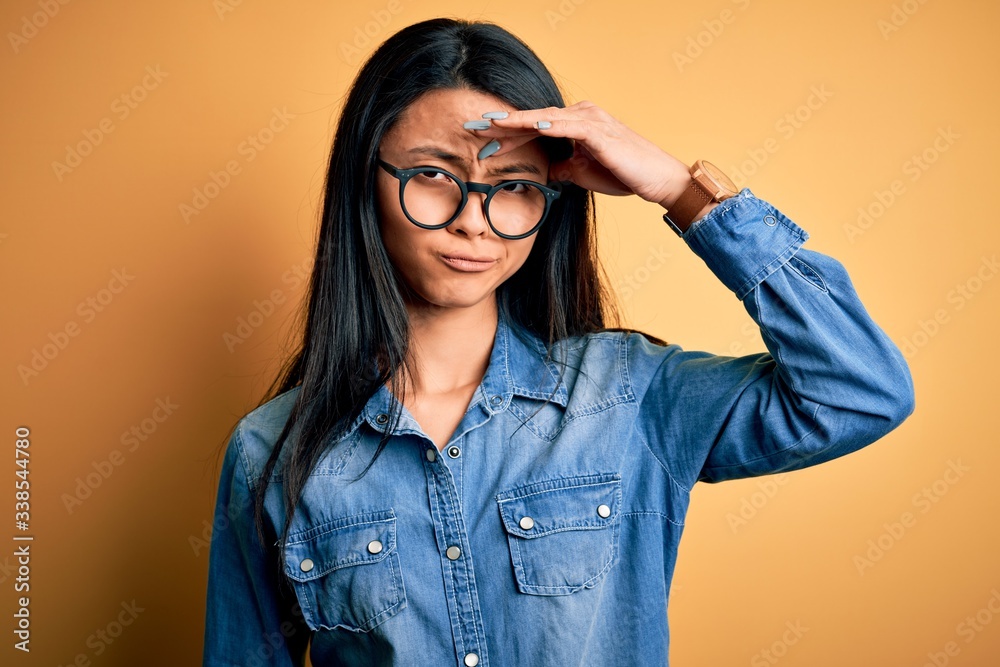Young beautiful chinese woman wearing casual denim shirt over isolated yellow background worried and stressed about a problem with hand on forehead, nervous and anxious for crisis