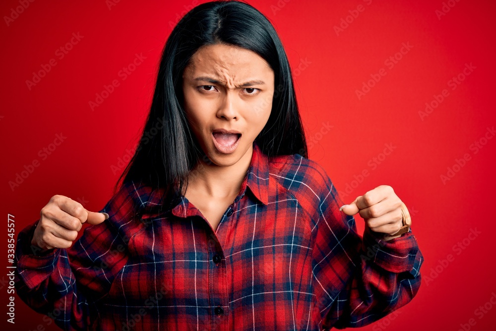 Young beautiful chinese woman wearing casual shirt over isolated red background angry and mad raising fists frustrated and furious while shouting with anger. Rage and aggressive concept. Young beautiful chinese woman wearing casual shirt over isolated red background angry and mad raising fists frustrated and furious while shouting with anger. Rage and aggressive concept.