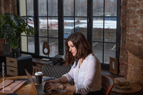 Business woman in white shirt working at laptop at home office near big window, drinking coffee. Chat on mobile. Wooden table, organizer.Remote work place. Quarantine of coronovirus pandemic covid-19