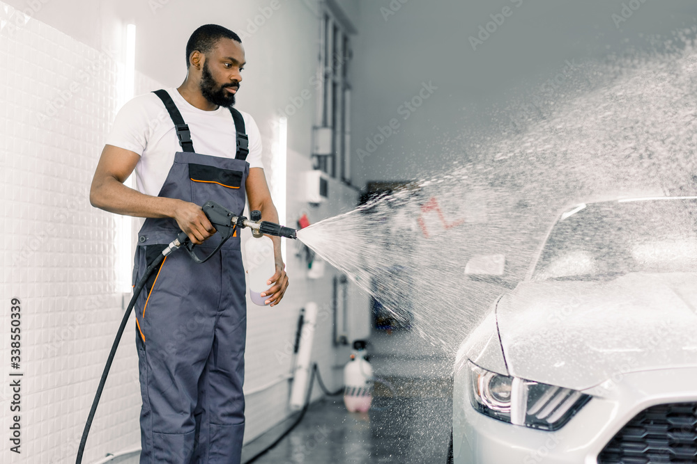 African American man, car wash worker is spraying cleaning foam to a modern  white luxury car holding a high pressure washer. Modern car and foam  washing, detailing wash foto de Stock