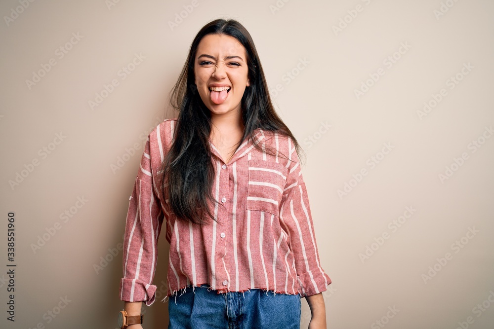 Young brunette woman wearing casual striped shirt over isolated background sticking tongue out happy with funny expression. Emotion concept. Young brunette woman wearing casual striped shirt over isolated background sticking tongue out happy with funny expression. Emotion concept.