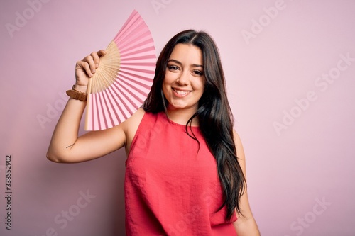 Young brunette woman holding hand fan for fresh air over pink isolated background with a happy face standing and smiling with a confident smile showing teeth © Krakenimages.com