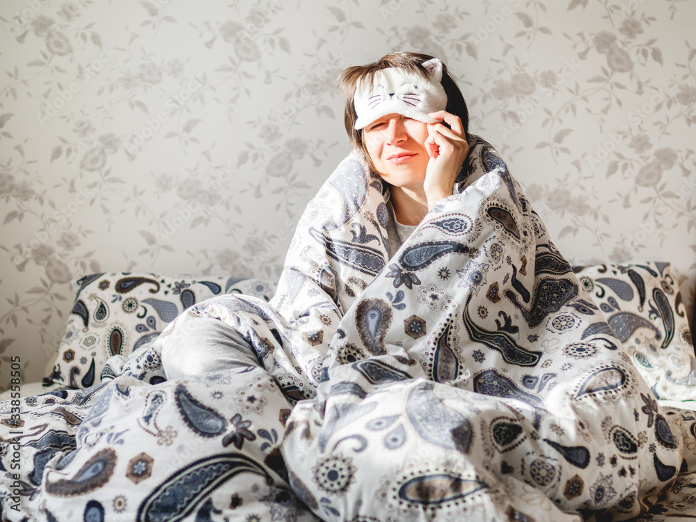 Young woman in grey pajama and sleeping mask in shape of cute sleeping cat face. She is just woke up and sit in bed. Early morning in cozy home. - obrazy, fototapety, plakaty 
