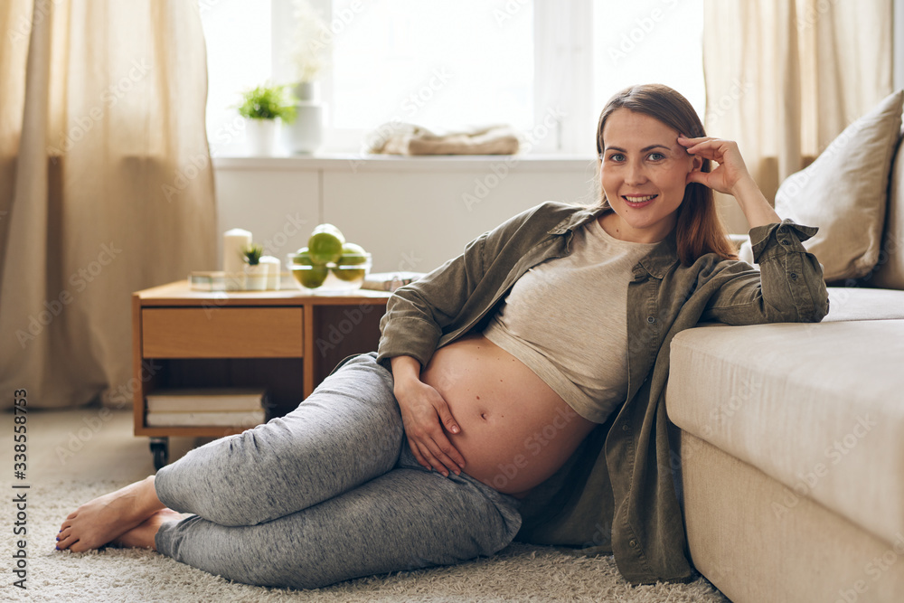 Portrait of smiling young pregnant woman sitting on floor and leaning on sofa while resting on maternity leave at home Portrait of smiling young pregnant woman sitting on floor and leaning on sofa while resting on maternity leave at home
