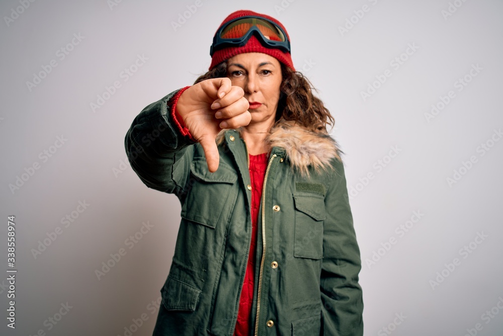 Middle age skier woman wearing snow sportswear and ski goggles over white background looking unhappy and angry showing rejection and negative with thumbs down gesture. Bad expression. Middle age skier woman wearing snow sportswear and ski goggles over white background looking unhappy and angry showing rejection and negative with thumbs down gesture. Bad expression.