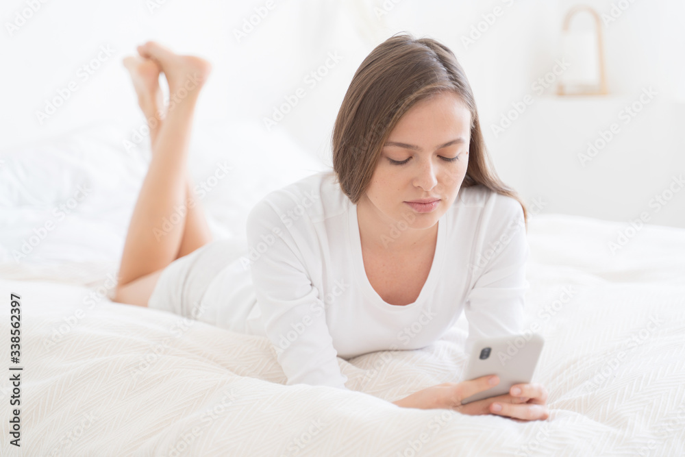 Young girl lying on bed, legs crossed, looking on smartphone screen she is holding in front, reading new messages or watching videos