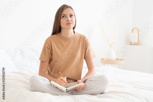 Young woman spending free time, sitting on bed in home clothes, writing down ideas and plans in her diary, willing to realize goals and dreams