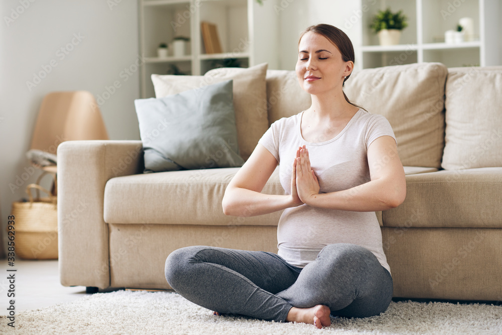 Content young pregnant woman sitting with hands in Namaste and meditating with closed eyes at home Content young pregnant woman sitting with hands in Namaste and meditating with closed eyes at home