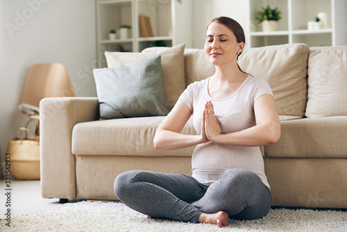 Content young pregnant woman sitting with hands in Namaste and meditating with closed eyes at home