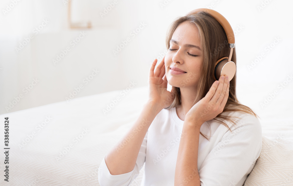 Girl in white top spending free time in bedroom in afternoon, enjoying favorite tracks through wireless headphones, keeping eyes closed, smiling Girl in white top spending free time in bedroom in afternoon, enjoying favorite tracks through wireless headphones, keeping eyes closed, smiling