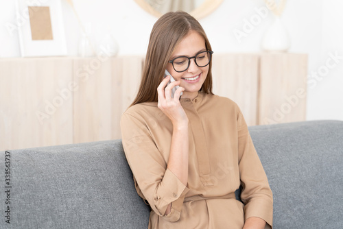 Young girl sitting on gray couch at home, wearing eyeglasses, talking on phone with friends discussing daily routine, feeling relaxed, smiling