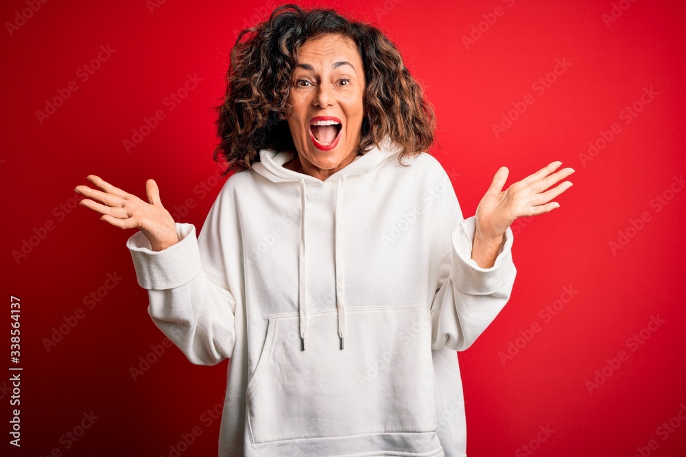 Middle age beautiful sporty woman wearing white sweatshirt over isolated red background celebrating crazy and amazed for success with arms raised and open eyes screaming excited. Winner concept Middle age beautiful sporty woman wearing white sweatshirt over isolated red background celebrating crazy and amazed for success with arms raised and open eyes screaming excited. Winner concept