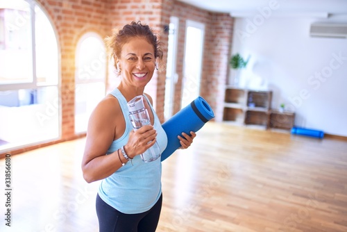 Middle age beautiful sportwoman smiling happy and confident. Standing with smile on face holding mat and bottle of water before doing exercise at gym photo