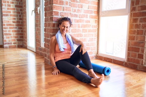 Middle age beautiful sportwoman smiling happy and confident. Sitting on the floor leaning on the wall with smile on face before doing exercise at gym