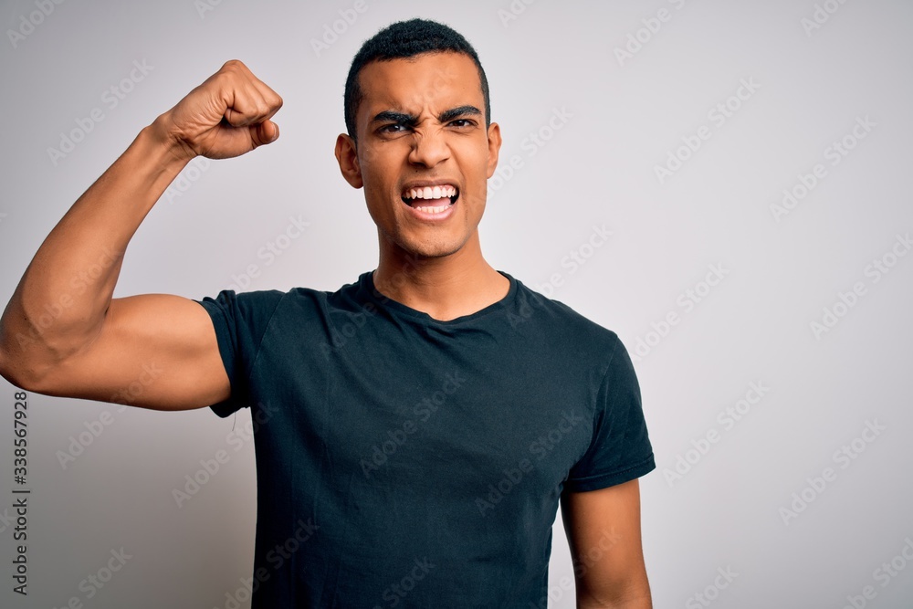 Young handsome african american man wearing casual t-shirt standing over white background angry and mad raising fist frustrated and furious while shouting with anger. Rage and aggressive concept.