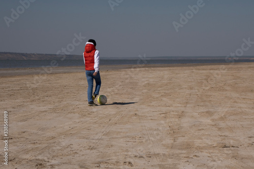 A boy of 10 years old in a white sweatshirt and orange vest plays football on a deserted beach in solitude.