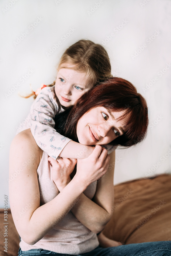 A little girl stands behind mom and hugs her with a pensive look. Young mother gently hugs daughter's hands. Close-up, light background. Parents and children. Mothers Day. Mom's love in everything A little girl stands behind mom and hugs her with a pensive look. Young mother gently hugs daughter's hands. Close-up, light background. Parents and children. Mothers Day. Mom's love in everything