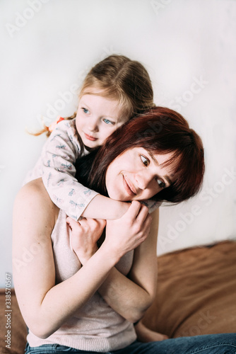 A little girl stands behind mom and hugs her with a pensive look. Young mother gently hugs daughter's hands. Close-up, light background. Parents and children. Mothers Day. Mom's love in everything