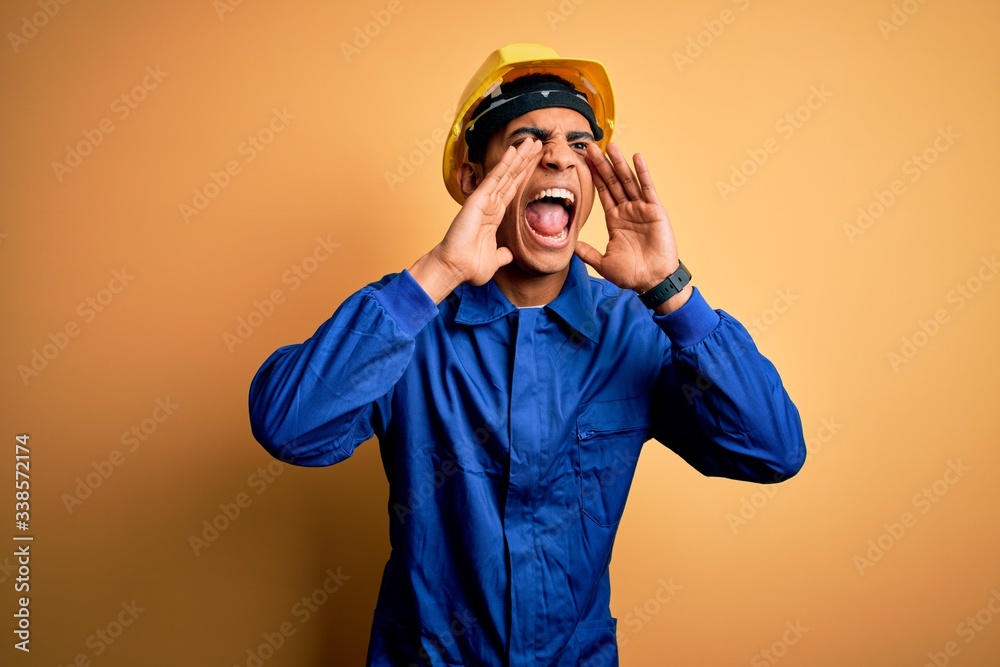Young handsome african american worker man wearing blue uniform and security helmet Shouting angry out loud with hands over mouth Young handsome african american worker man wearing blue uniform and security helmet Shouting angry out loud with hands over mouth
