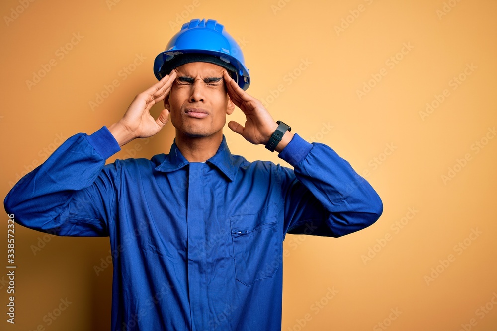 Young handsome african american worker man wearing blue uniform and security helmet suffering from headache desperate and stressed because pain and migraine. Hands on head.